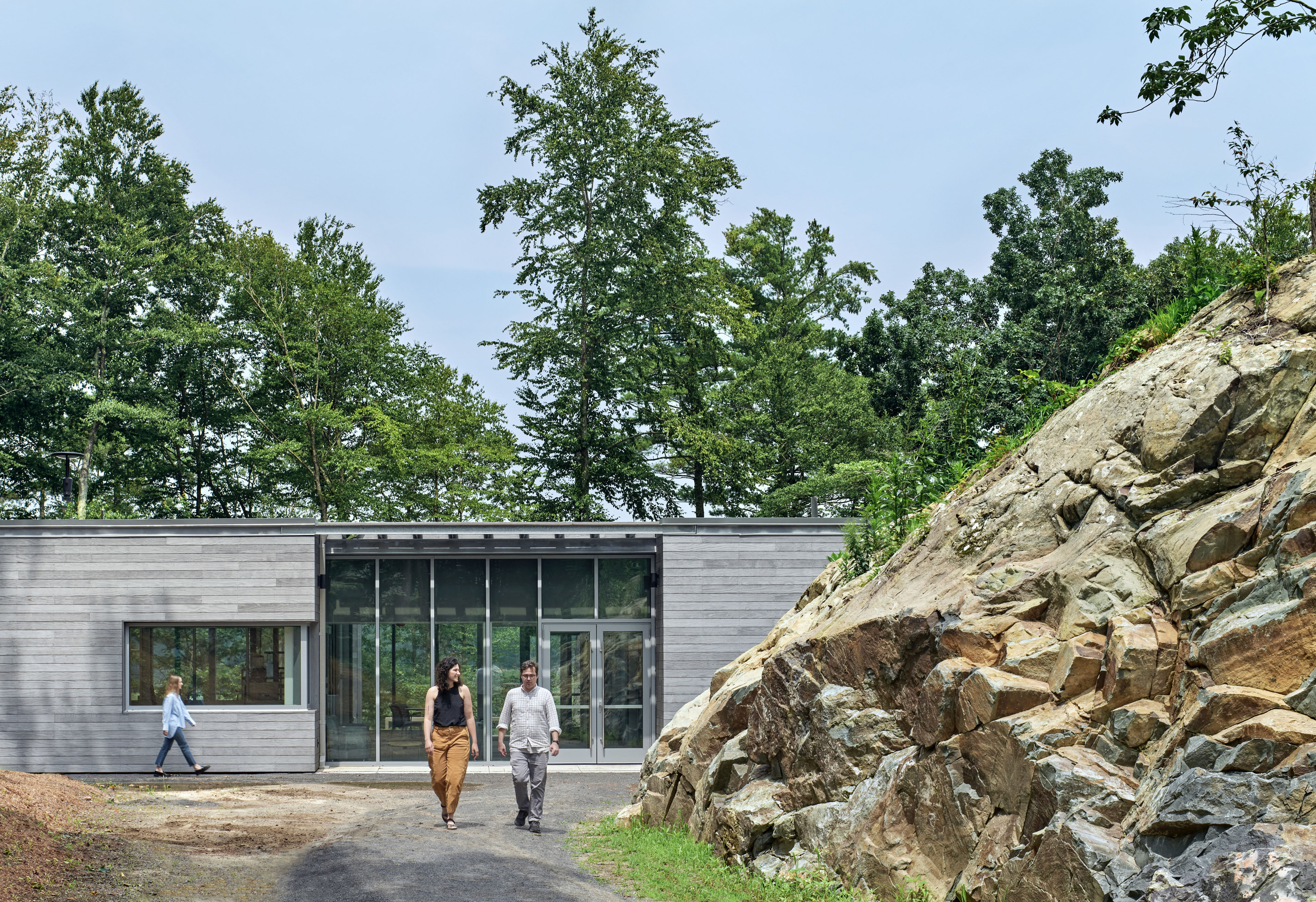 Walkway of Alfond Research Center with vertical wood on left and right sides with 6 vertical windows in entry way. Forest fills back of image and large rock of formation sits to the right