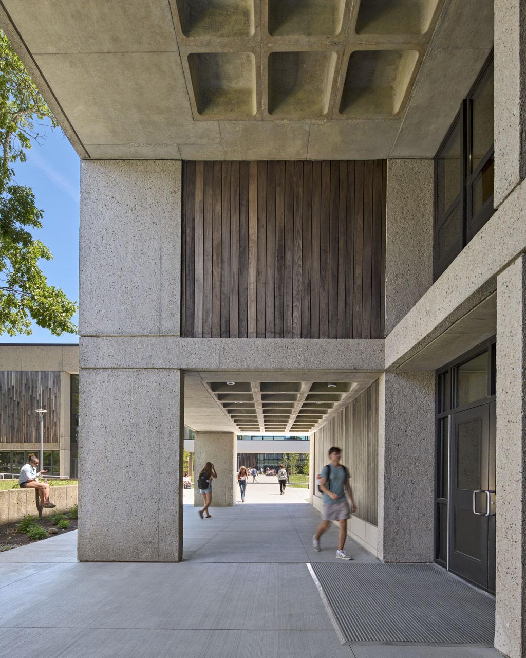Students walking down outdoor corridor with covered top with varying heights, additional buildings connecting on the left