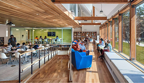 Students reading in classroom with bench lounge seating to the right with windows looking out to lawn, walkway in the middle, and desks to the left