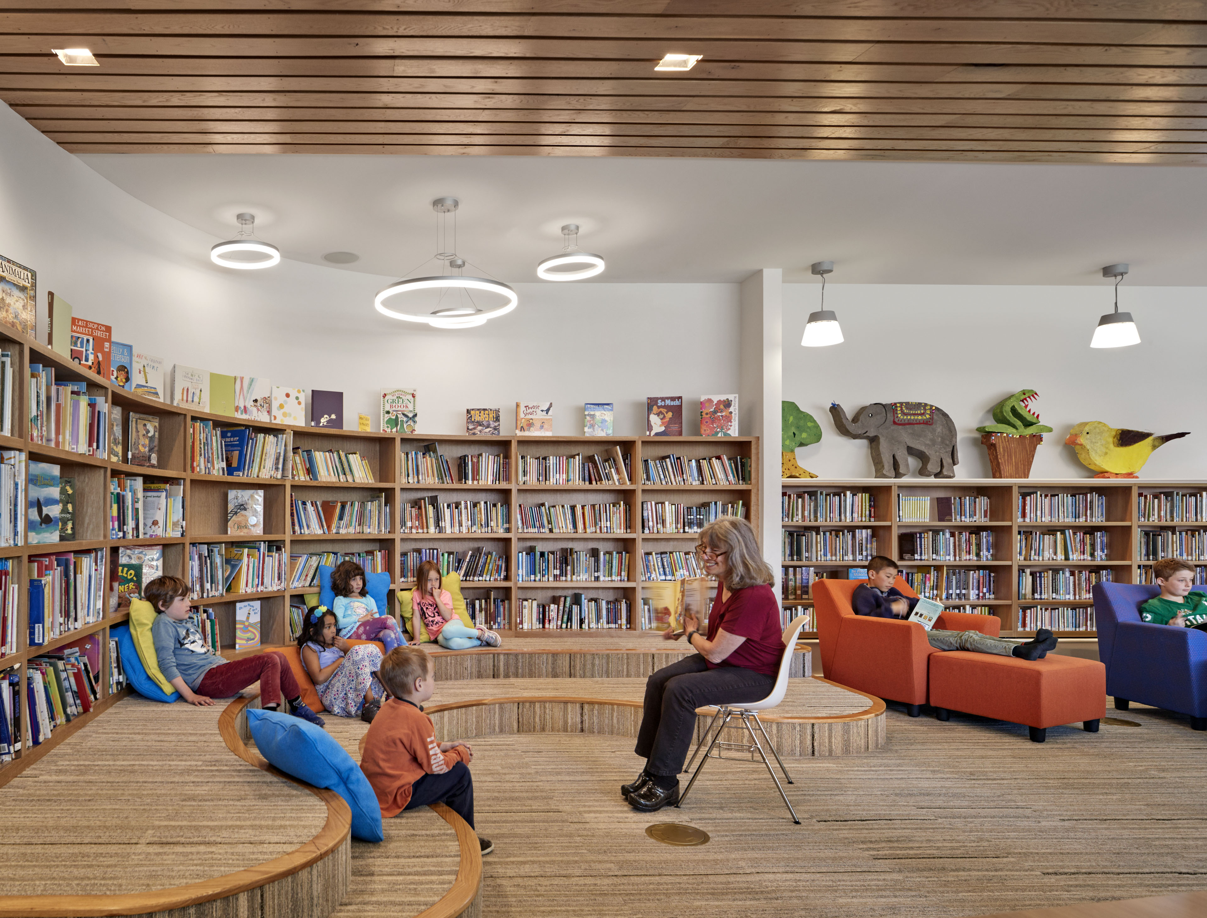 Library with curved staggered seating area with students, librarian in chair reading to students, books lining the side walls