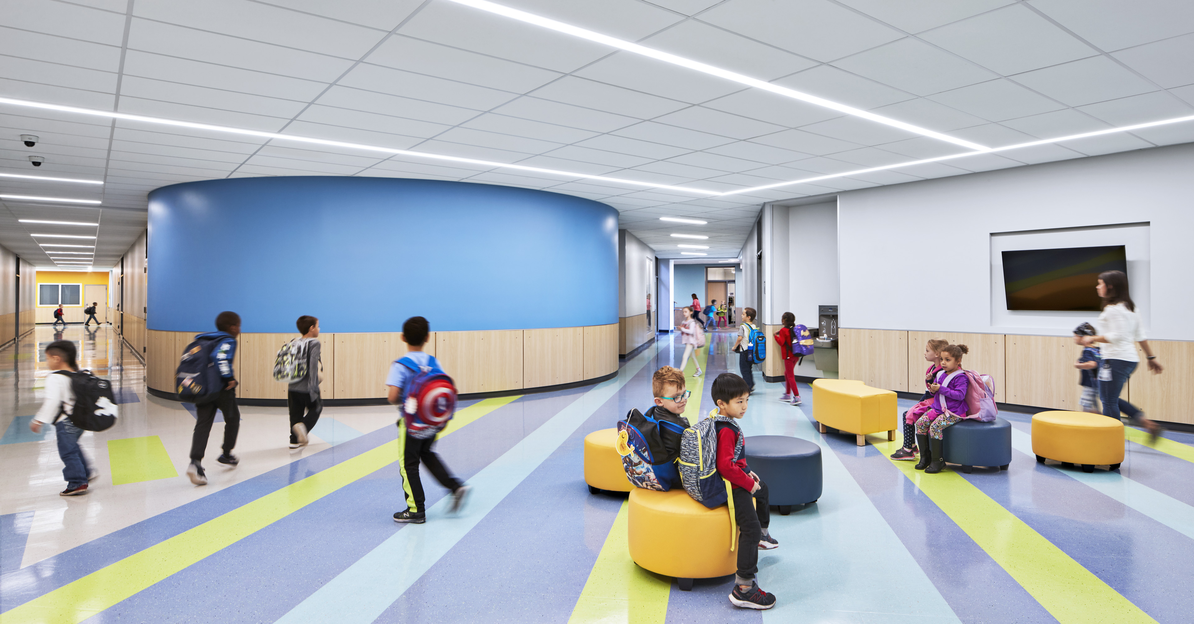 Interior with lime green, dark and light blue stripes with yellow and blue ottomans and kids seated and standing. Hallways to left and right with blue circular wall in middle with wood paneling the bottom half