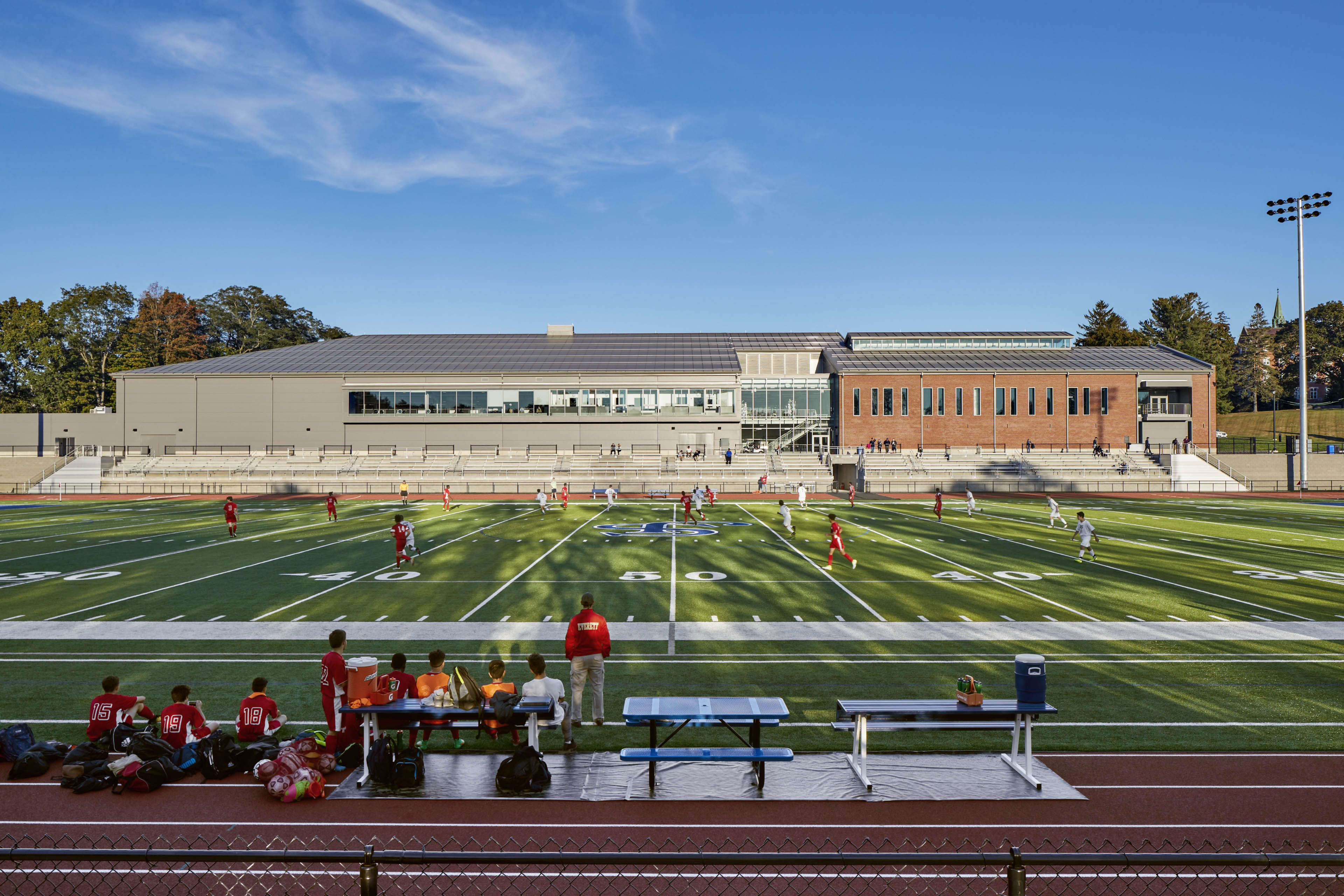 exterior of school with students practicing on football field, stadium seating behind and light shining on building
