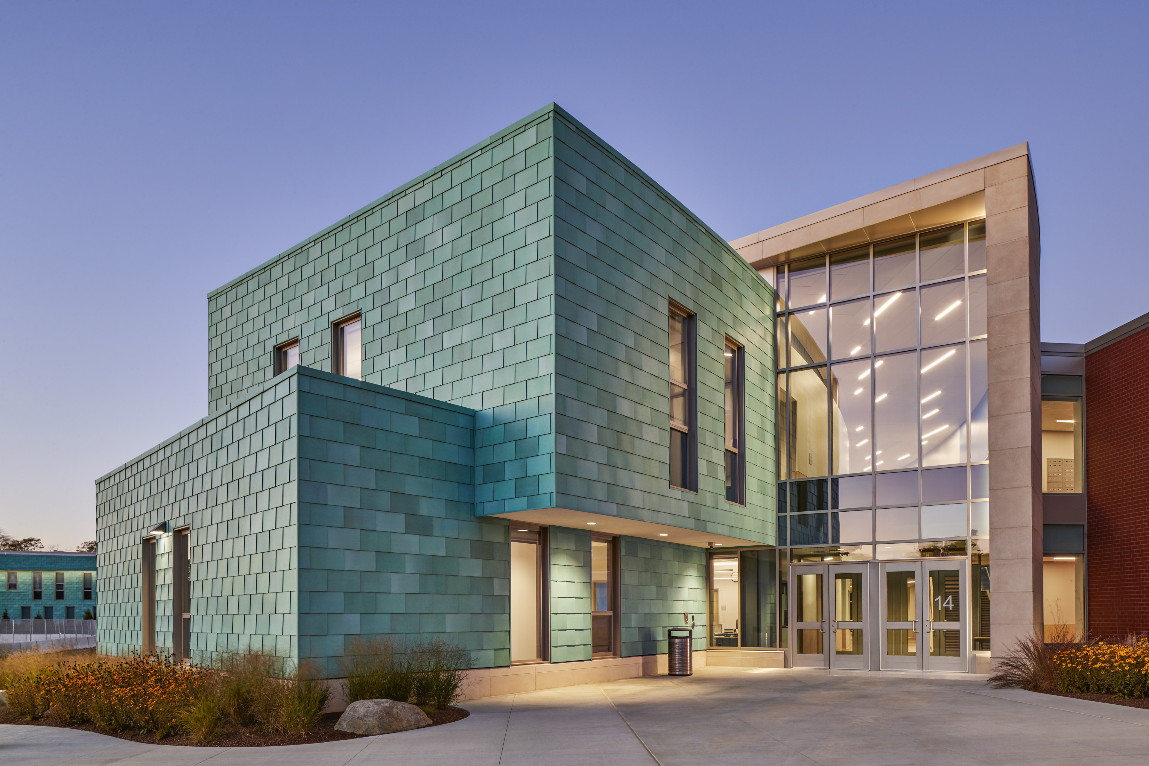 Entrance with stacked appearance with aqua tiles, and large vertical windows on unit with doors to the right. Purple and blue sky and small bushes surrond