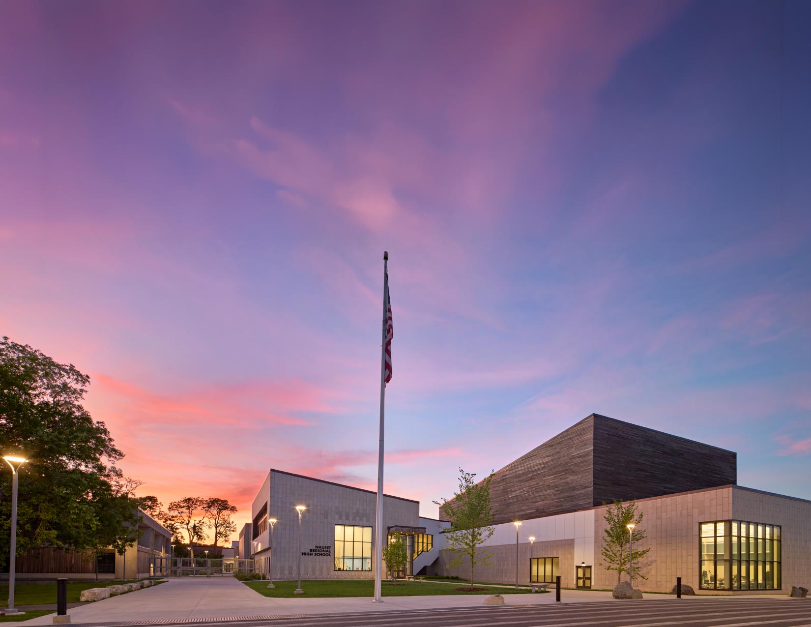 Entrance of school with pink and purple sunset draping the sky. Flagpole in center of image with amber glow from classrooms