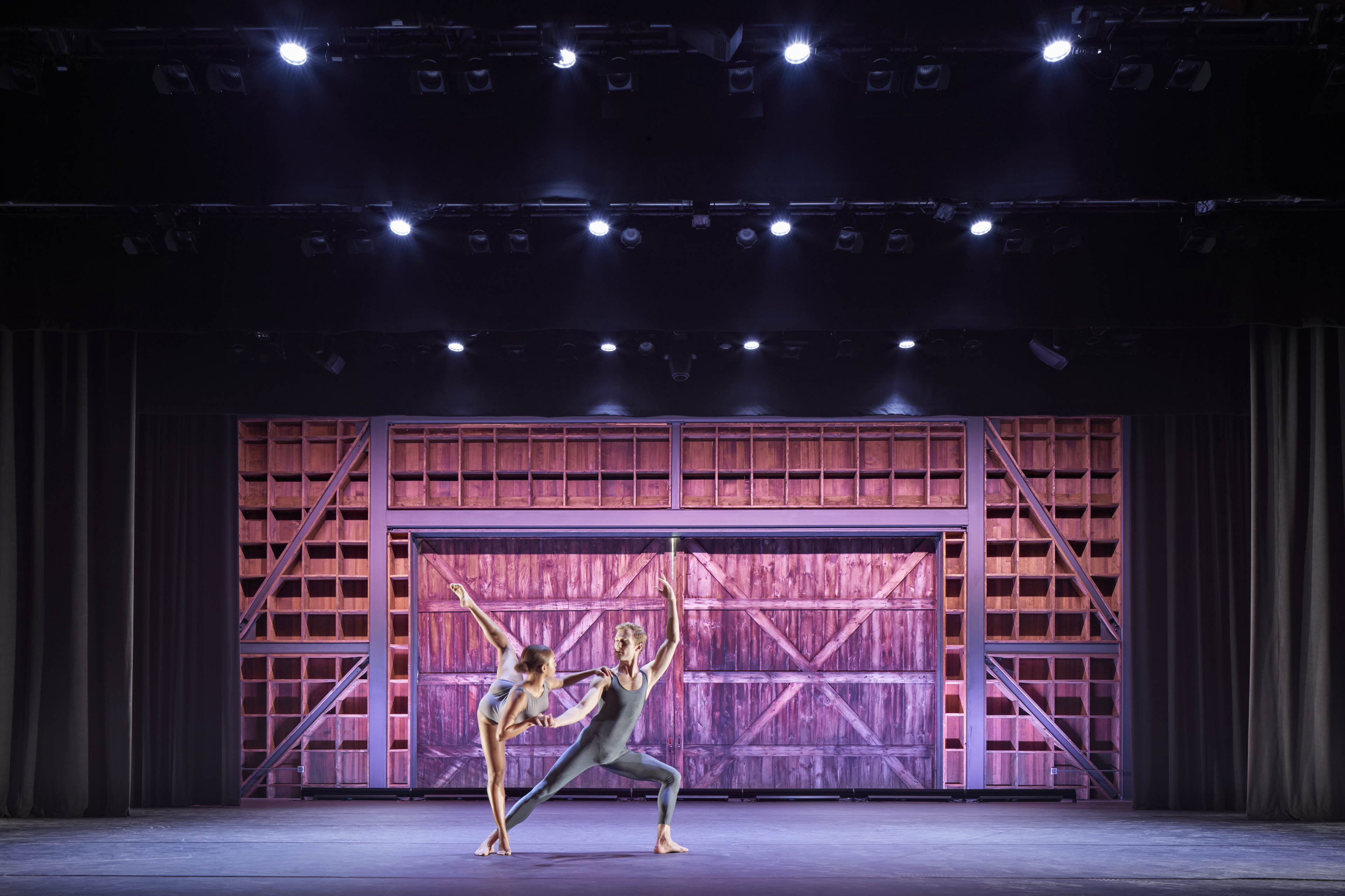 ballerinas on stage and wood in small square formation with barn door to the right and pink lighting displayed across backgrond