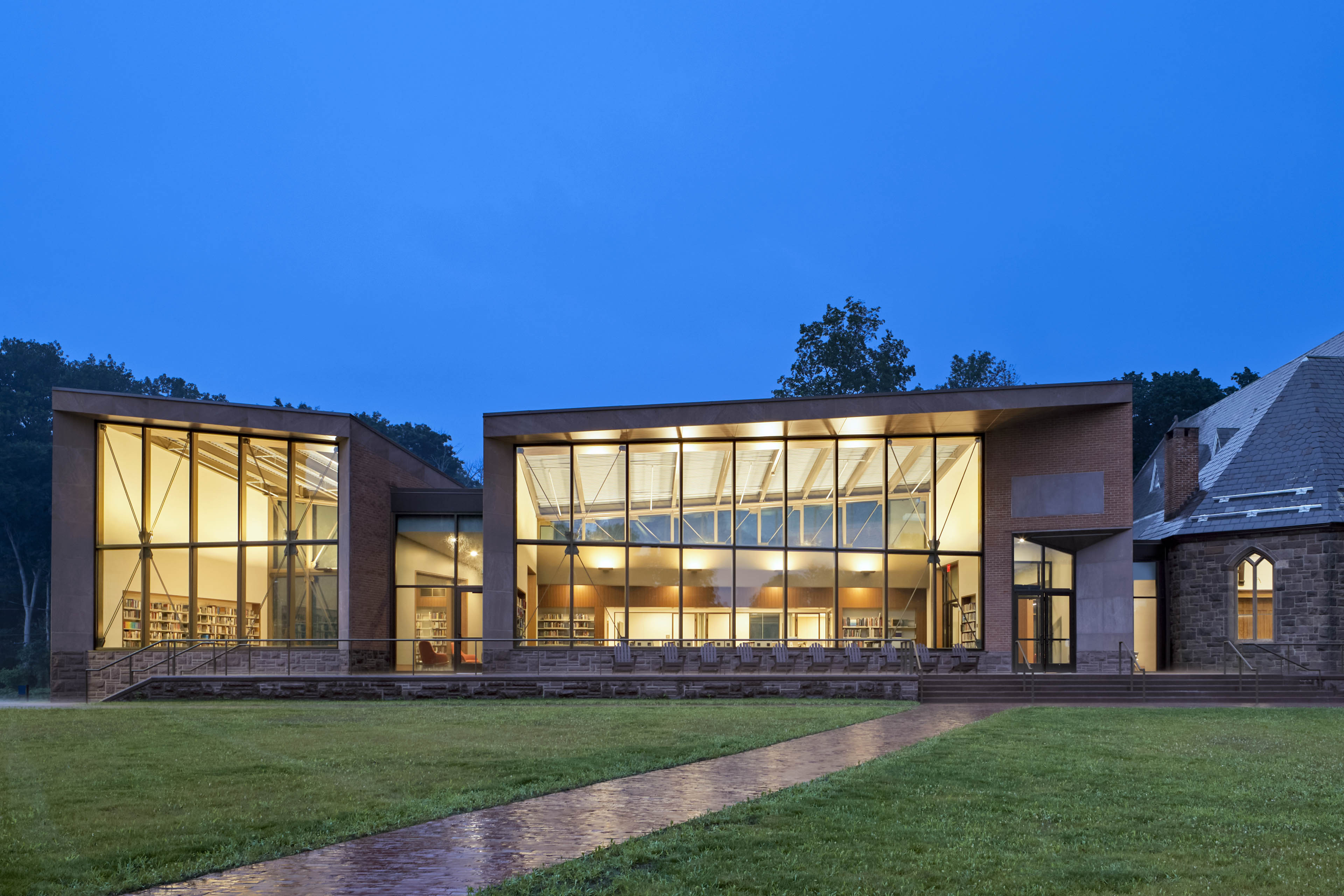 View of exterior of building at night with yellow light glowing out. Walkway in front with grass on both sides
